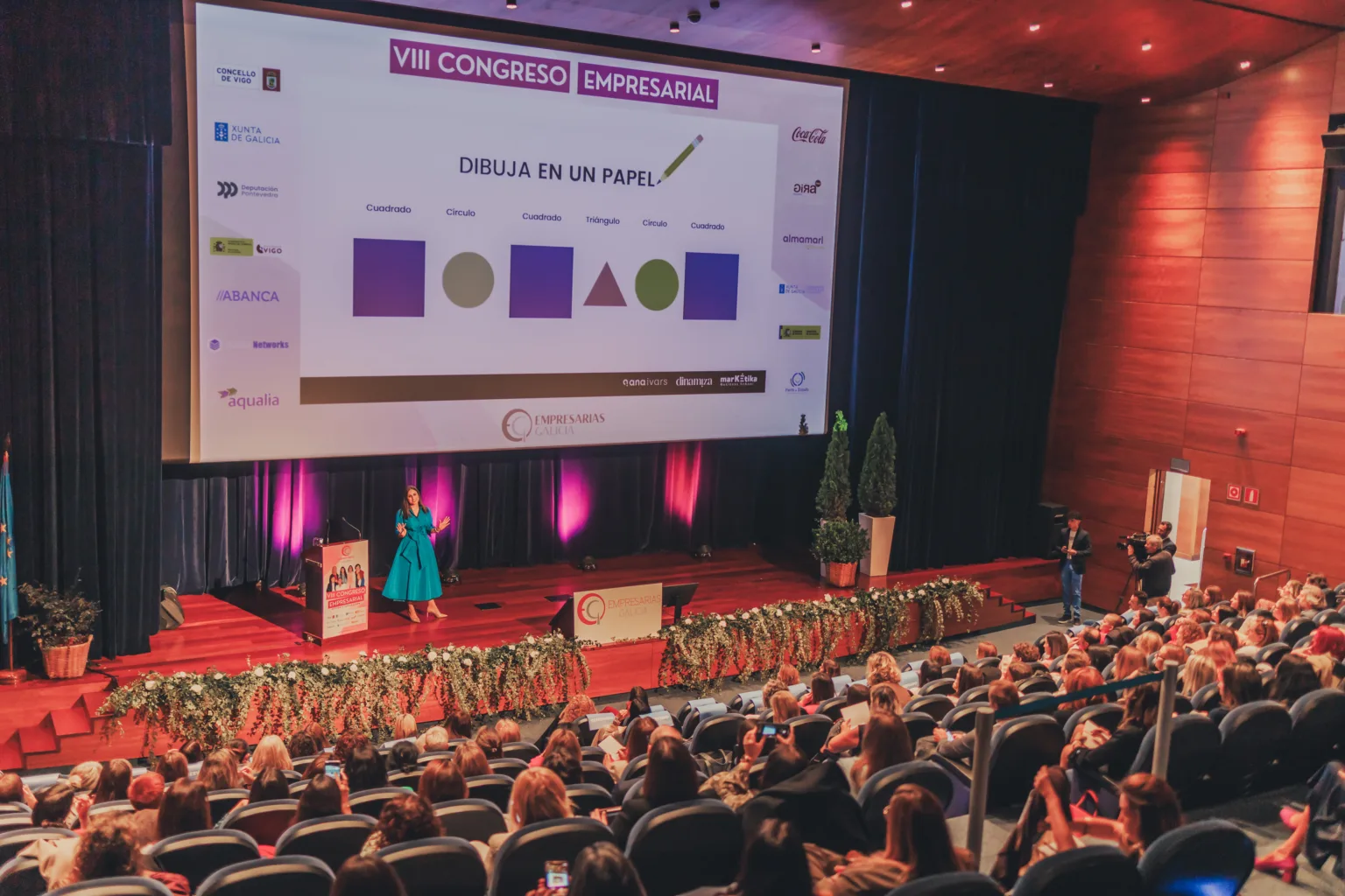 Fotografía para eventos empresariales en el VIII Congreso Empresarial de Empresarios Galicia, con Ana Ivars, consultora de marketing digital, en el escenario hablando al público. Incluye asistentes atentos, una cámara de televisión grabando y la pantalla del proyector desplegado.