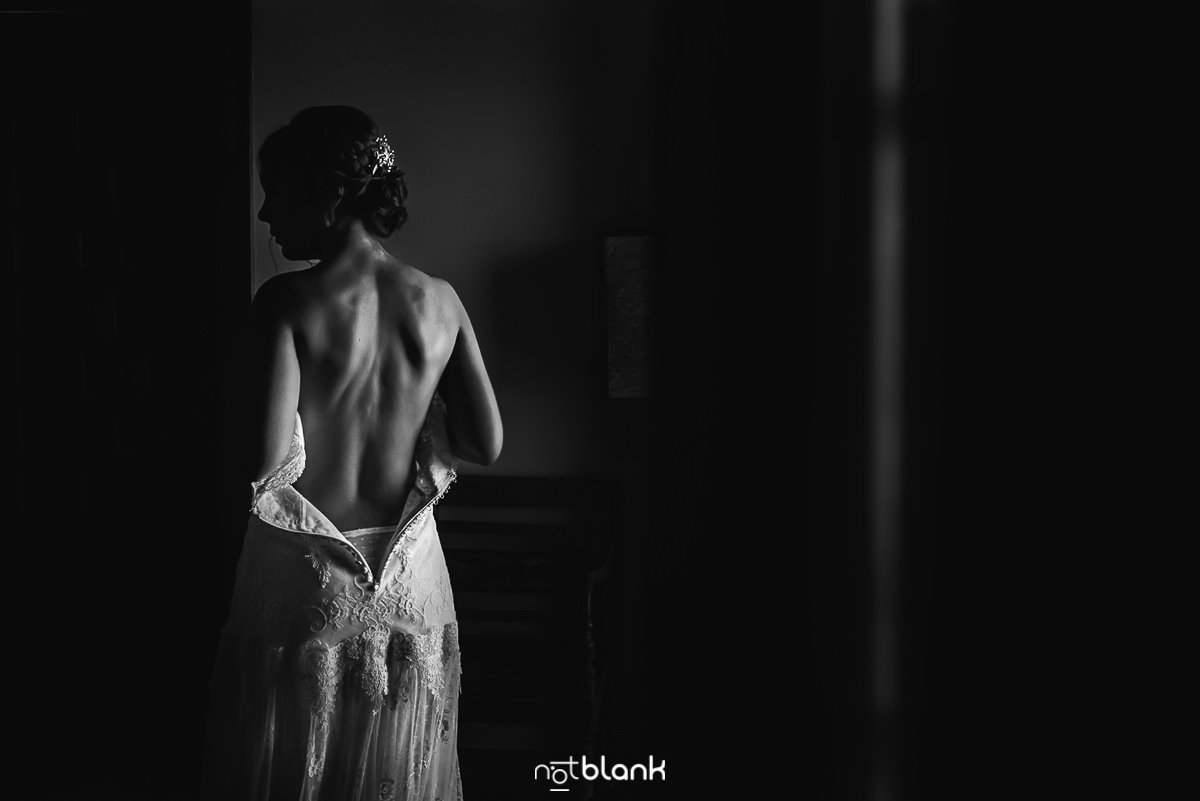 Novia de espaldas junto a una ventana, con luz natural iluminando su espalda y el contorno de sus brazos en una habitación oscura.