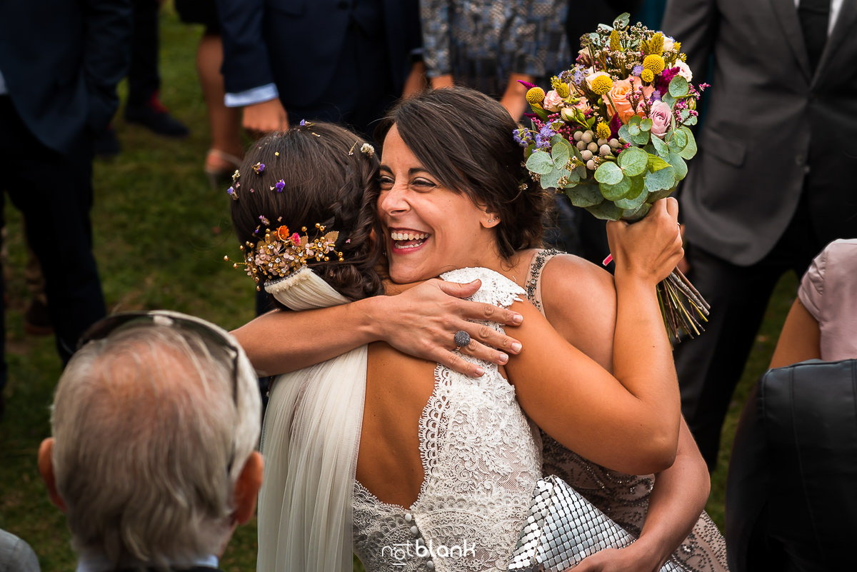 "Vista desde arriba de la novia abrazando a una invitada justo después de la ceremonia. La novia, de espaldas, muestra una gran sonrisa que refleja la alegría del momento, mientras los invitados permanecen fuera de foco en el fondo.