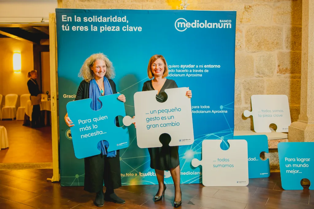 Dos consultoras del Banco Mediolanum posan frente a un photocall con el branding de la entidad durante un evento solidario en Casona La Torre, Redondela. Sostienen piezas de puzzle gigantes que forman frases como “Para quien más lo necesita, un pequeño gesto es un gran cambio”, reflejando el eslogan del evento: “En la solidaridad, tú eres la pieza clave”