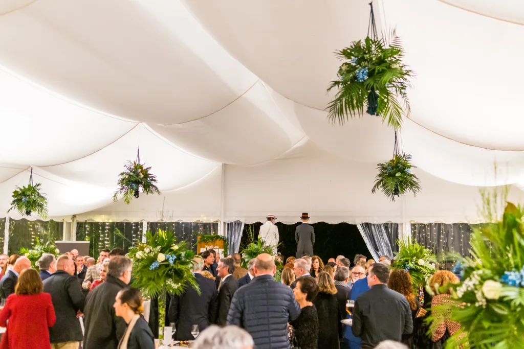 Panorámica de la carpa decorada para el 25 aniversario de Aemos, mostrando las cabezas de los invitados bajo un techo blanco con ondas y plantas colgantes. Al fondo, dos zancudos con trajes blanco y gris animan el evento, reflejando la atención al detalle en esta celebración empresarial.