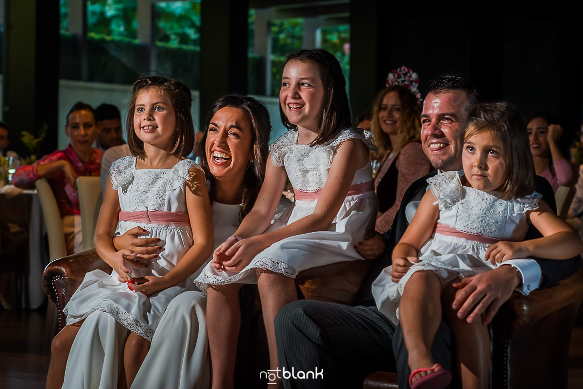 Los novios y sus tres sobrinas reaccionan al same day edit, proyectado el día de la boda. La novia y el novio muestran una gran carcajada, contagiando la alegría a dos de sus sobrinas, mientras en el fondo se intuyen los invitados desenfocados.