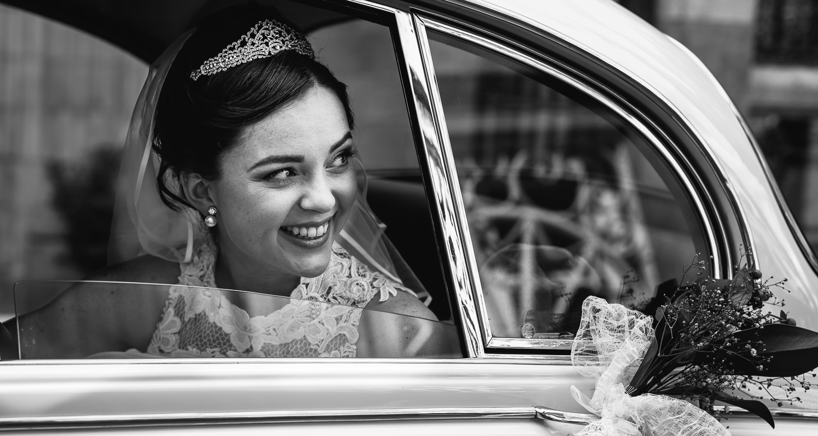 Novia sonriendo, luciendo una pequeña corona, dentro de un Jaguar clásico, mirando hacia los invitados en la entrada de la iglesia.