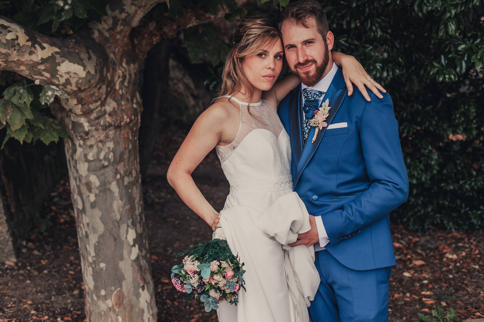 Novios posando bajo la sombra de un árbol. La novia sostiene un ramo y tiene una mirada penetrante; el novio, con traje azul, le acaricia suavemente el vestido mientras sonríe levemente.