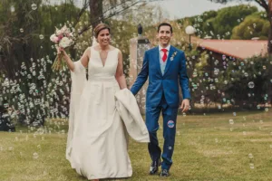 Pareja caminando de la mano en una finca, sonrientes y rodeados de pompas de jabón, listos para sorprender a los invitados en su boda