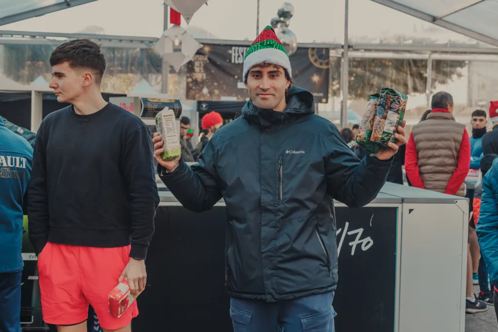 Fotografía para eventos solidarios en Galicia: Manu Ochoa, deportista olímpico de Tomiño, sonriente con gorro de Papá Noel y paquetes de comida no perecedera durante la Carrera Solidaria San Silvestre 2024.