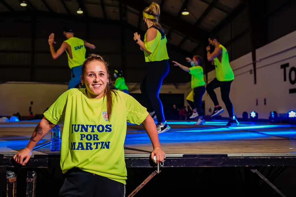 Fotografía para eventos solidarios en Galicia: Nuria en el escenario del evento solidario de fundraising presencial Juntos por Martín, con chicas bailando zumba en el fondo.