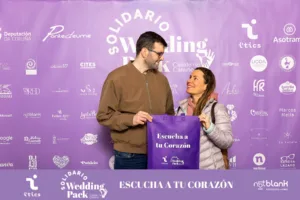 Pareja visitante del Wedding Pack Solidario viéndose entre ellos y sujetando una bolsa con el lema "Escucha tu corazón" frente al photocall del evento.